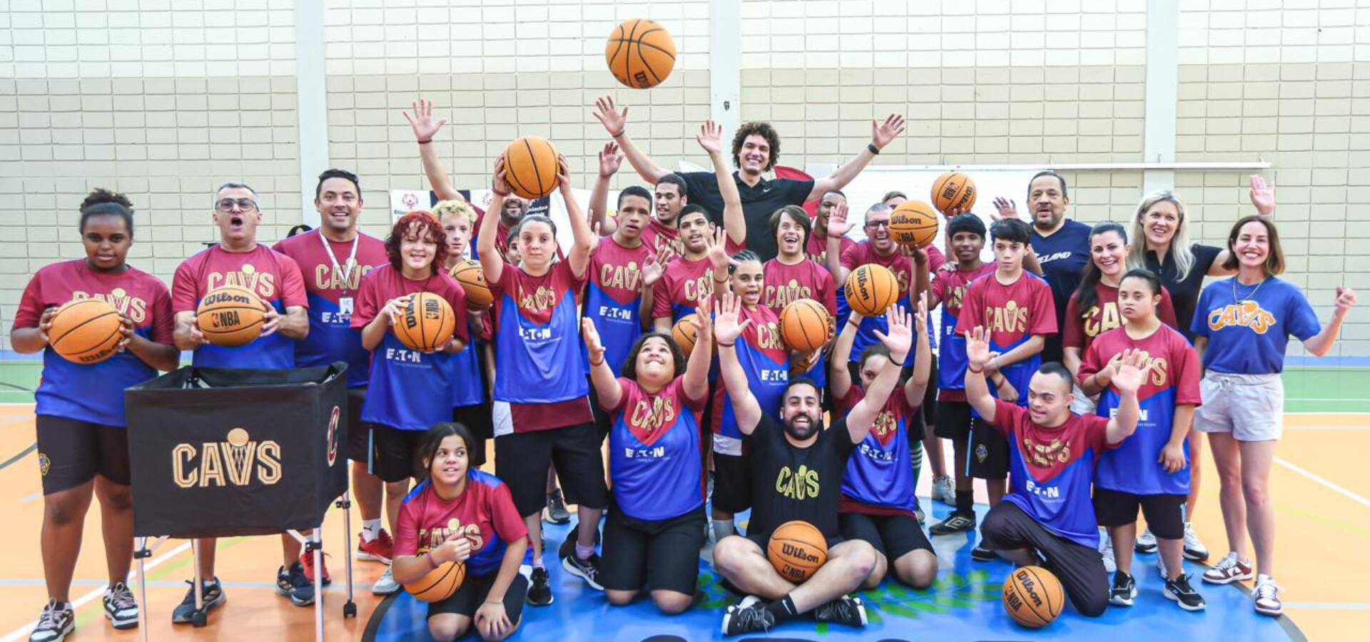O embaixador da OEB, Anderson Varejão, usa uma camiseta preta e se encontra no centro da imagem, atrás dos atletas.
